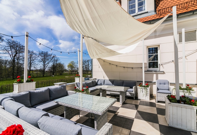Słoneczny taras hotelowy z szarą plecioną sofą narożną, szklanymi stolikami, żaglem przeciwsłonecznym, lampkami i czerwonymi kwiatami
