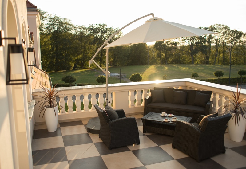 Słoneczny taras hotelowy z wiklinową sofą i fotelami, parasolem, filiżankami na stoliku i widokiem na park