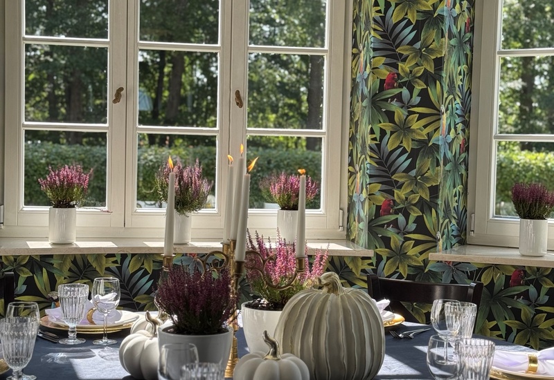 Stół przy dużych oknach: czarny obrus, białe dekoracyjne dynie, wrzosy, świecznik i talerze ze złotą obwódką