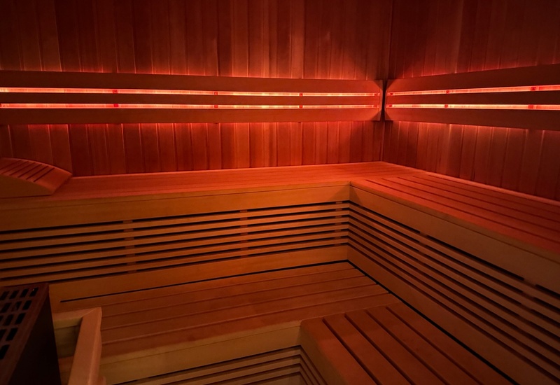 Sauna w hotelu z drewnianymi, wielopoziomowymi ławami narożnymi, zagłówkiem i ciepłym czerwonym oświetleniem LED.