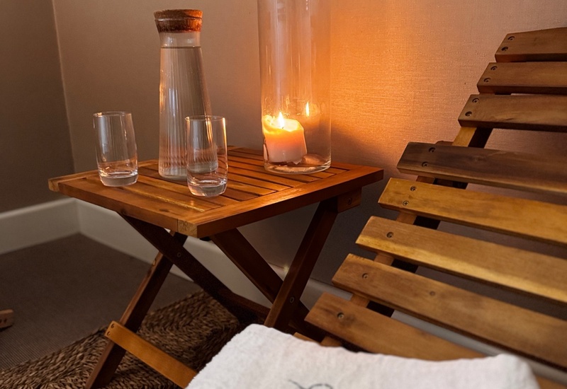 Drewniany leżak i stolik z karafką wody, dwiema szklankami, palącą się świecą w szkle i białym haftowanym ręcznikiem