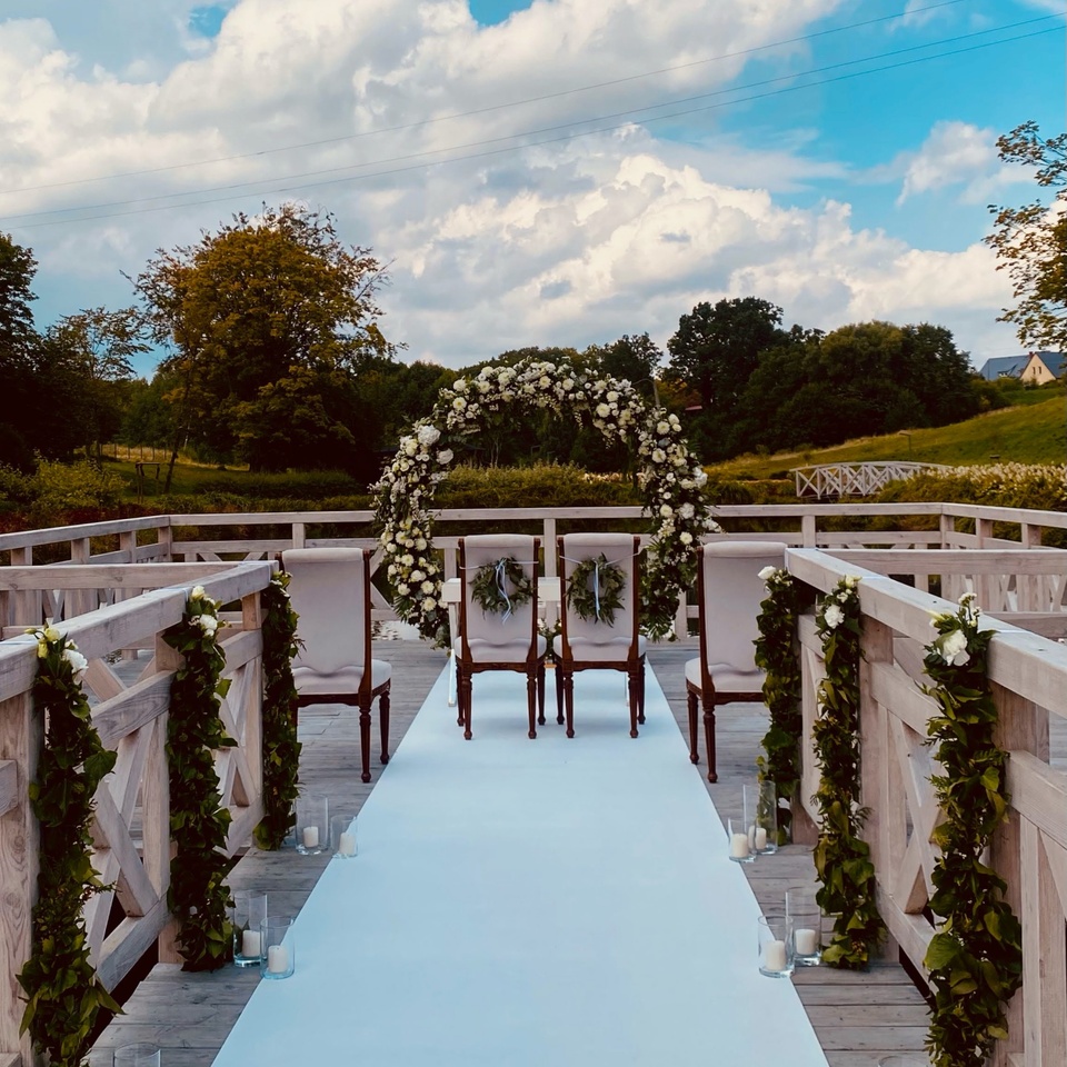 Drewniany pomost nad jeziorem na ceremonię ślubną z białym dywanem, kwiatowym łukiem, ozdobnymi krzesłami i świecami.