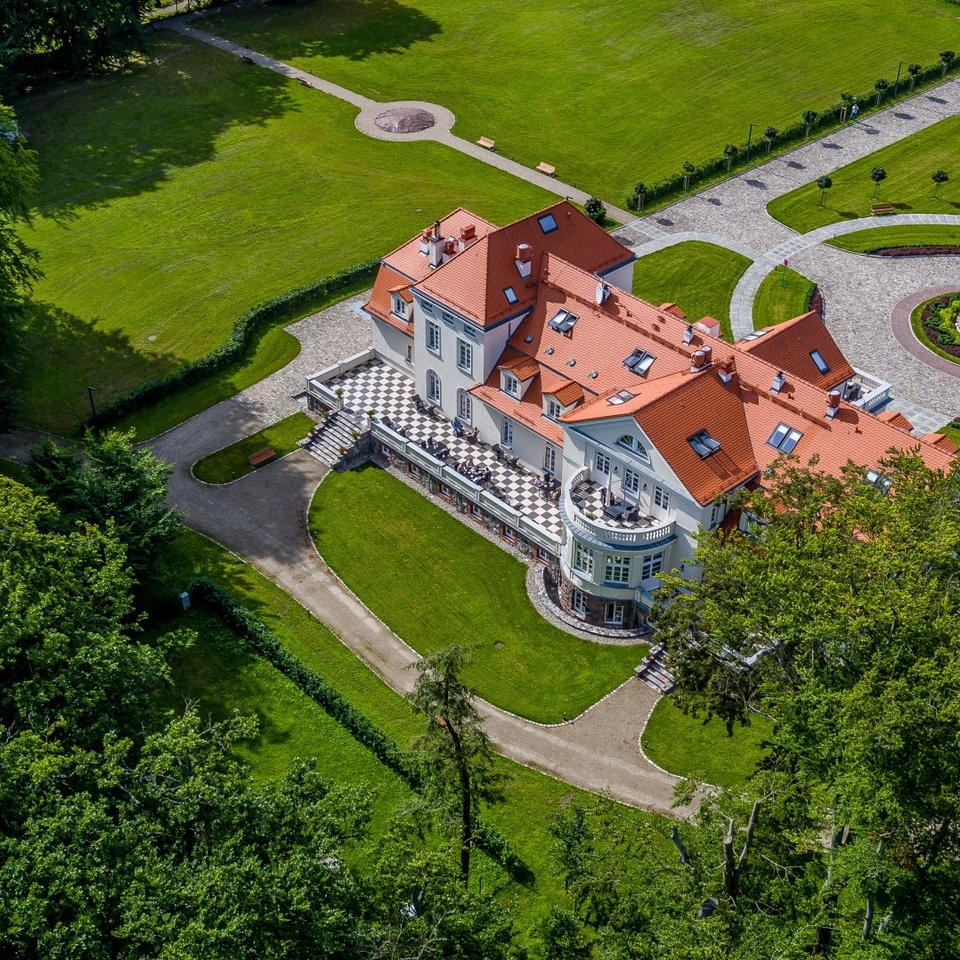 Hotel butikowy z czerwoną dachówką, tarasem w szachownicę, okrągłym podjazdem z fontanną oraz zadbanymi trawnikami i parkiem.