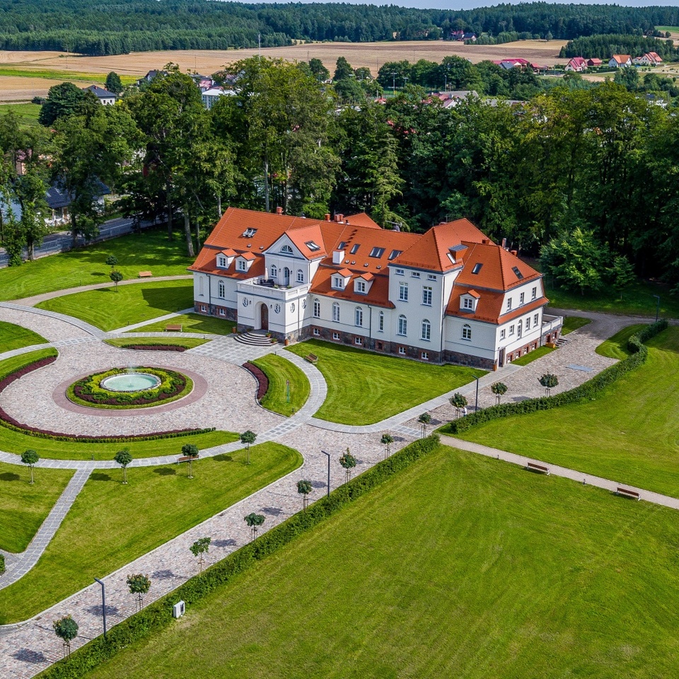 Pałacyk hotelowy w stylu manor z czerwonym dachem, brukowanym okrągłym dziedzińcem z fontanną, zadbanymi trawnikami, parkingiem i zalesieniem w tle