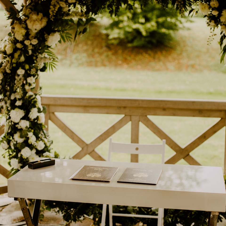 Biały stół do podpisów z dwoma dokumentami pod bujną kwiatową arkadą w drewnianej altanie, plener ślubny