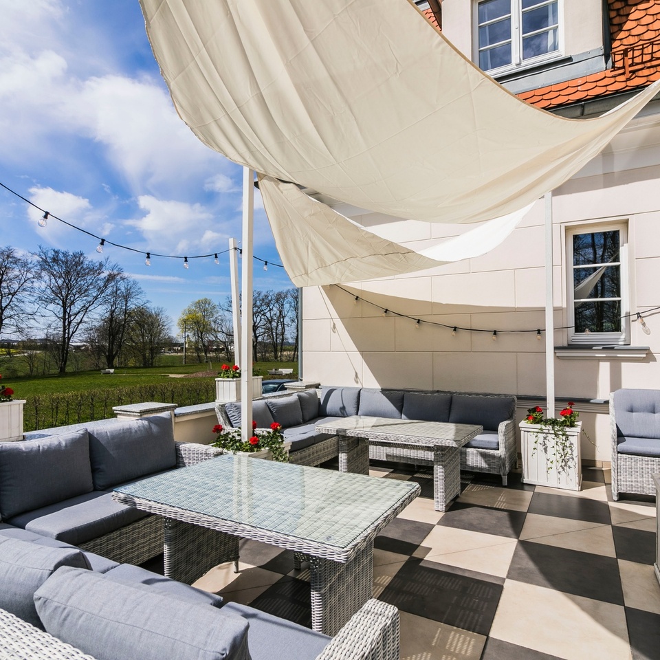 Słoneczny taras hotelowy z szarą plecioną sofą narożną, szklanymi stolikami, żaglem przeciwsłonecznym, lampkami i czerwonymi kwiatami