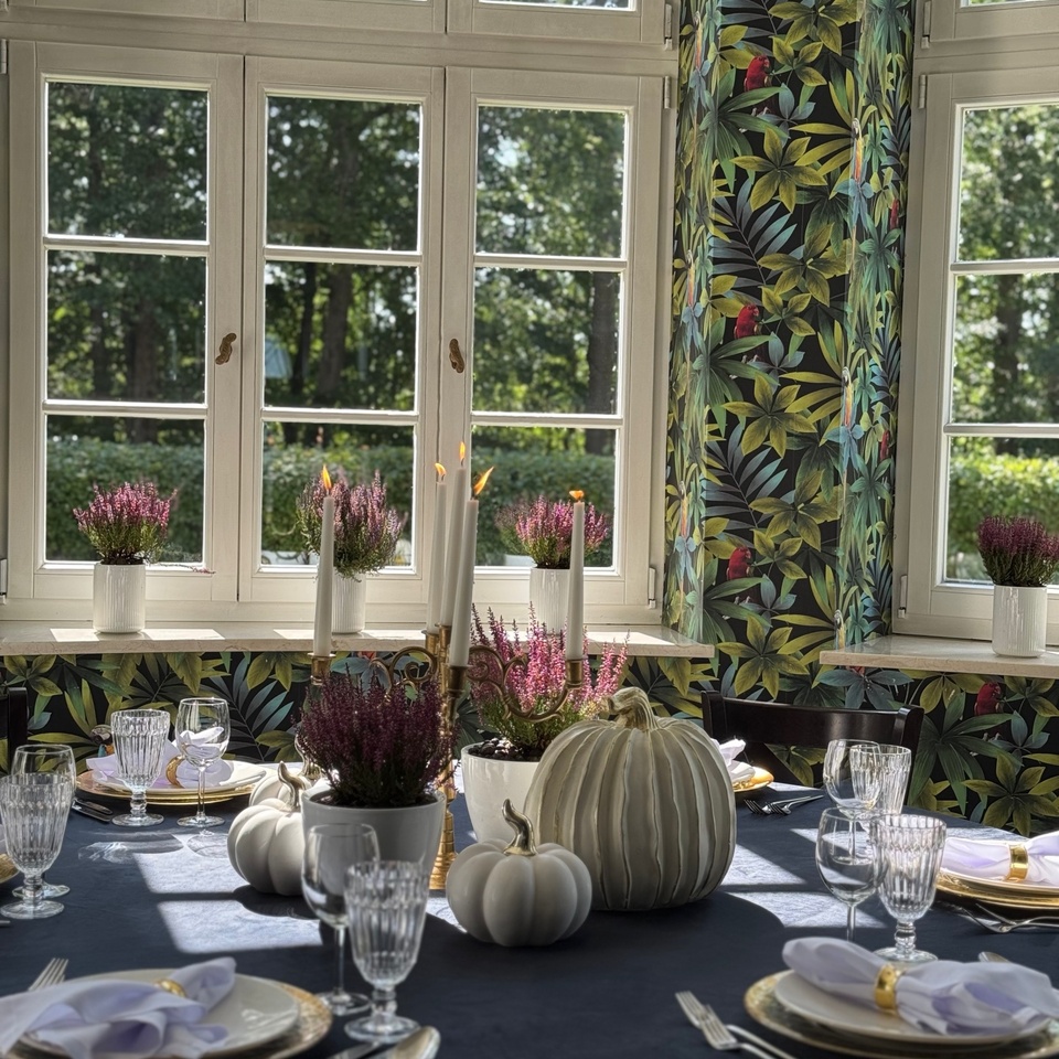 Stół przy dużych oknach: czarny obrus, białe dekoracyjne dynie, wrzosy, świecznik i talerze ze złotą obwódką