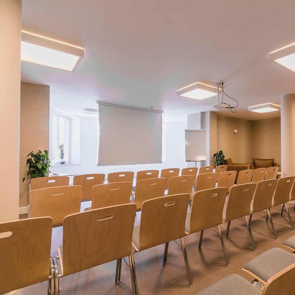 Jasna nowoczesna sala konferencyjna z rzędami drewnianych krzeseł, projektorem sufitowym, ekranem rozwijanym, flipchartem i roślinami.