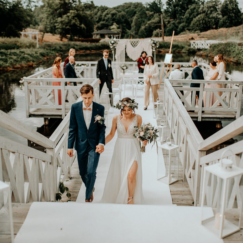Para młoda idzie za rękę po białym drewnianym pomoście nad jeziorem, z makramowym łukiem, gośćmi i latarniami