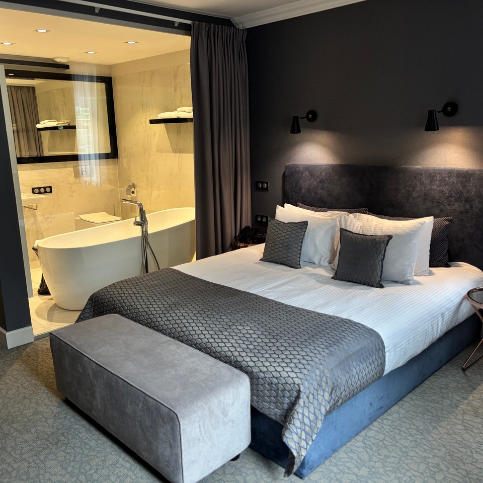 Nowoczesny pokój z łóżkiem typu king, miękkim zagłówkiem, lampkami nocnymi, pufą i wolnostojącą wanną za przeszkloną ścianką