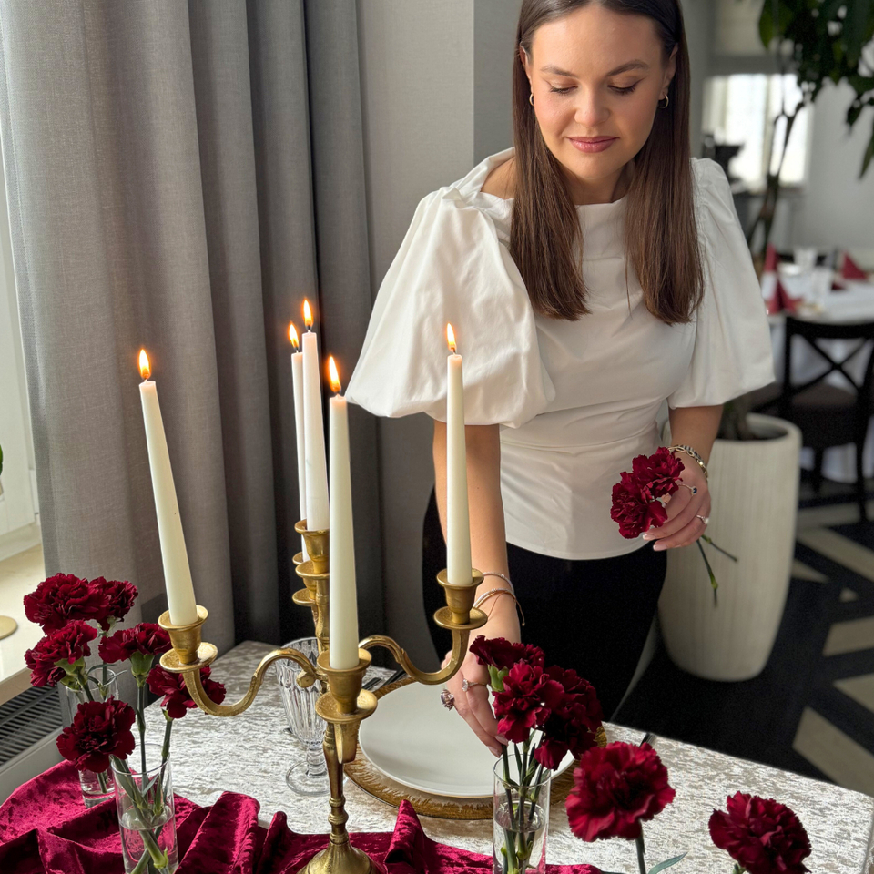 Kobieta ustawia elegancki stół z palącym świecznikiem, czerwonymi goździkami, aksamitnym bieżnikiem i złotymi talerzami.