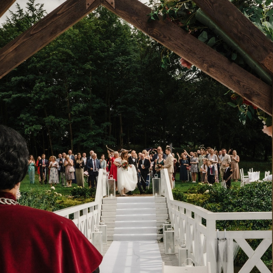 Plenerowy ślub na białym moście z drewnianą arkadą ozdobioną kwiatami, para młoda schodzi alejką, goście wiwatują.