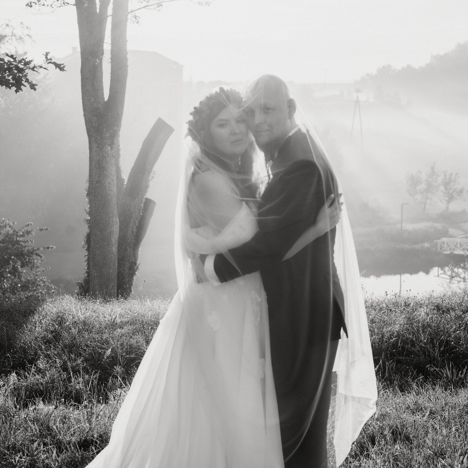 Romantyczne, jeziorne zdjęcie ślubne o porannej mgle: panna młoda w zwiewnej sukni i welonie przytula się do pana młodego na hotelowym trawniku.