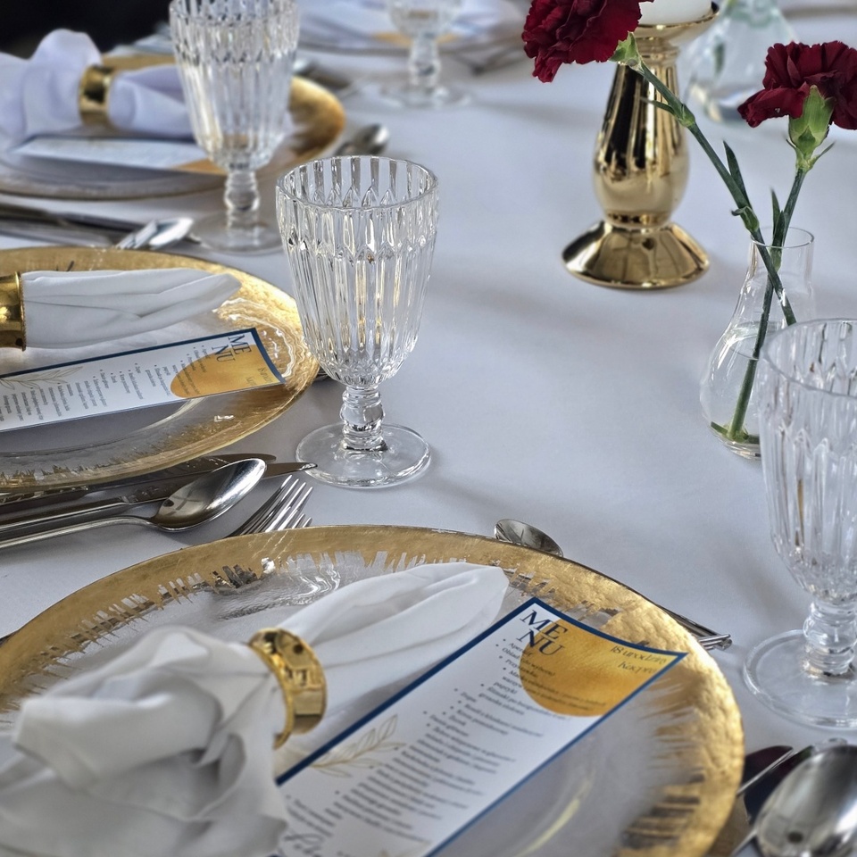 Eleganckie nakrycie stołu: złote talerze, kryształowe kielichy, białe serwety w złotych obrączkach, menu i czerwone goździki.