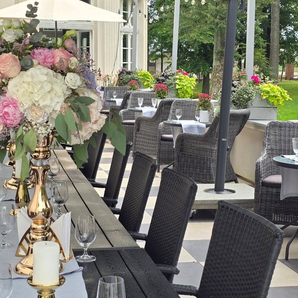 Taras hotelowy z długim ciemnym stołem, złotymi świecznikami, bukietem kwiatów, fotelami rattanowymi i widokiem na ogród
