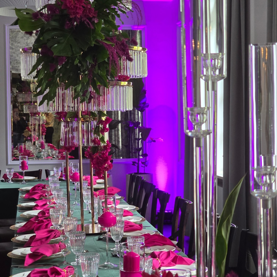 Długi stół bankietowy z zielonym obrusem, fuksjowymi serwetkami, szkłem i wysokimi kwiatami w fioletowym świetle