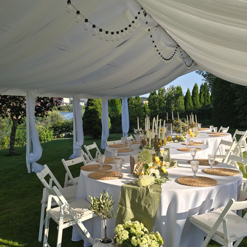 Biały, drapowany namiot z girlandą świateł nad okrągłymi stołami w białych obrusach, podkładkami, wysokimi świecami, kwiatami i koszami z cytrynami