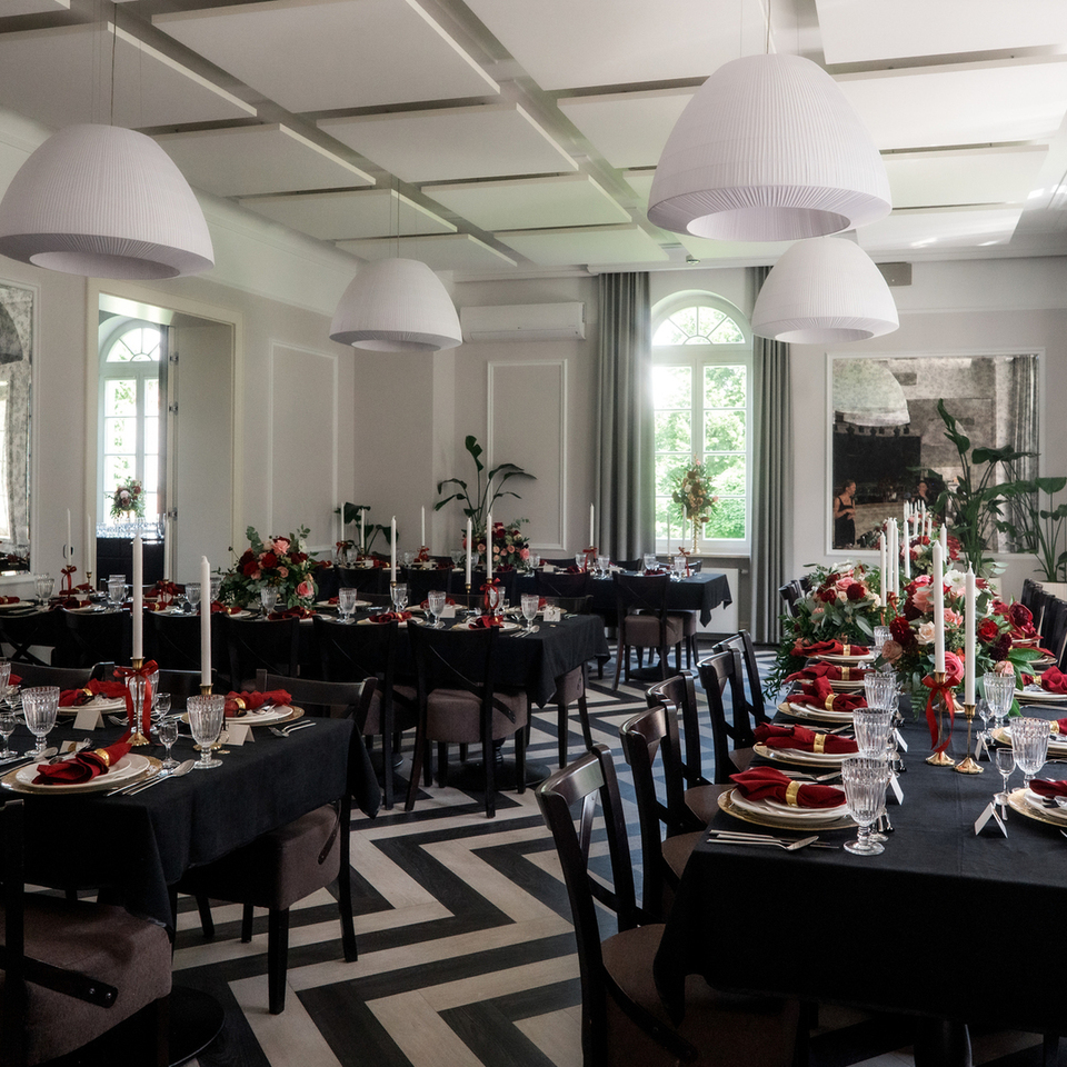Elegancka sala bankietowa z długimi stołami w czarnych obrusach, czerwonymi serwetkami, kwiatowymi aranżacjami, wysokimi świecami, dużymi lampami i półokrągłymi oknami