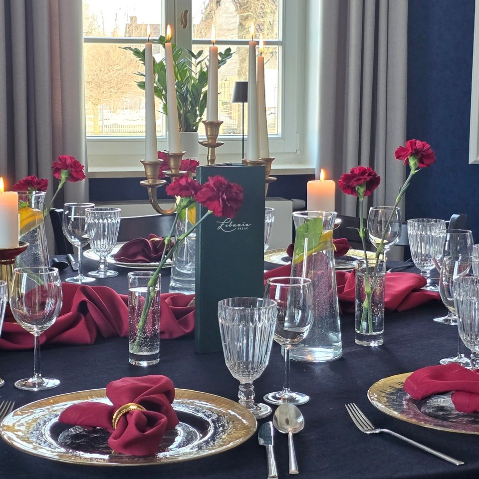 Elegancki hotelowy stół nakryty czarnym obrusem, złote talerze, bordowe serwety w złotych pierścieniach, kryształowe szkło, świeczki i czerwone goździki.