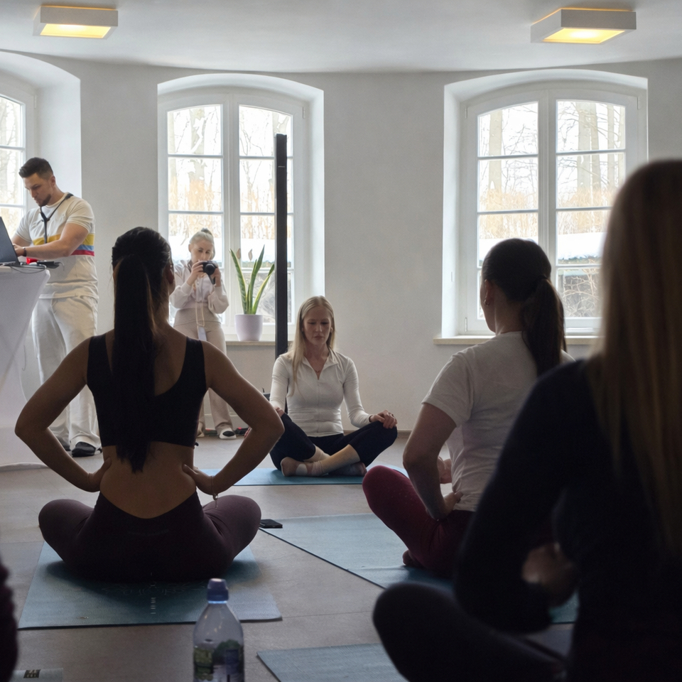 Studio jogi w hotelu z łukowymi oknami; instruktorka medytuje z przodu, goście na matach, łagodne światło i butelka wody