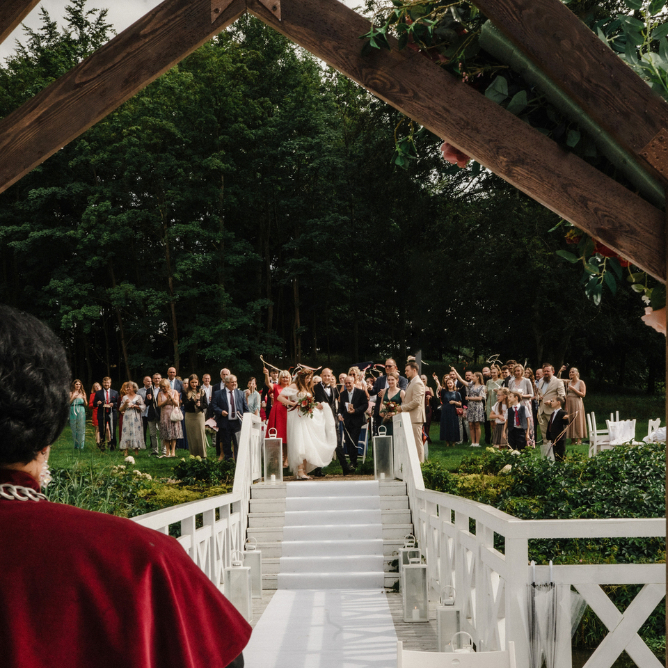 Weselna ceremonia w ogrodzie pod drewnianą, kwiatową arkadą; biały dywan na moście, para młoda przywitana przez gości.