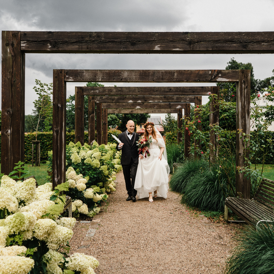 Para młoda idzie pod drewnianymi pergolami aleją obsadzoną hortensjami, z bukietem, zadbane trawniki i ławki pod pochmurnym niebem