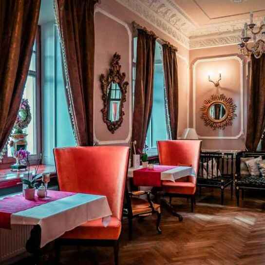 Przytulny, elegancki salonik hotelowy z wysokimi różowymi fotelami, kryształowym żyrandolem i lustrami.