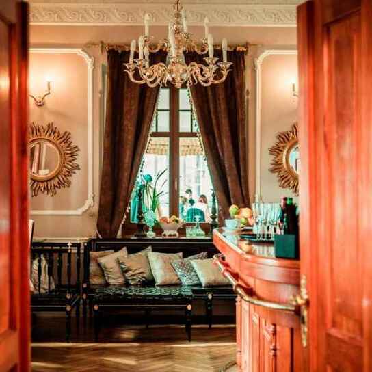 Wejście do eleganckiego hotelowego salonu z drewnianymi drzwiami, kryształowym żyrandolem, ozdobnymi lustrami, kanapą z poduszkami i barem.