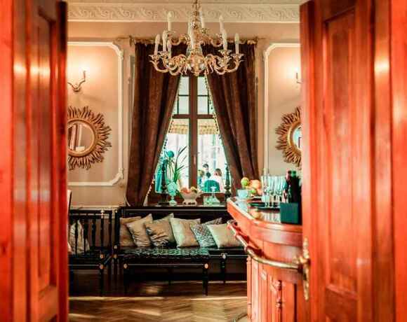 Wejście do eleganckiego hotelowego salonu z drewnianymi drzwiami, kryształowym żyrandolem, ozdobnymi lustrami, kanapą z poduszkami i barem.