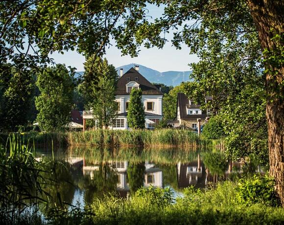 Urokliwy hotel z tarasem nad jeziorem, biały willowy budynek odbija się w wodzie, otoczony drzewami i górami.