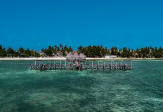 Overwater wooden pier with thatched-roof huts extending into clear turquoise ocean near a sandy beach lined with palm trees