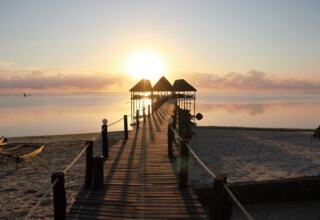 Wooden pier with thatched-roof pavilions stretches over calm beach waters at sunrise, offering serene waterfront views