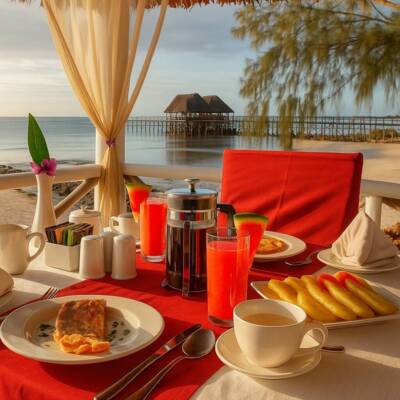Beachfront breakfast setup with red chairs, fresh juice, coffee, pastries, and ocean view featuring overwater bungalows at sunrise