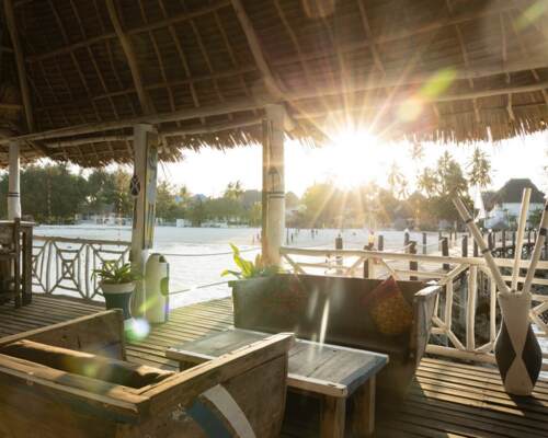 Sunlit rustic beachside lounge with wooden furniture, decorative cushions, potted plants, and a view of sandy shore and palm trees