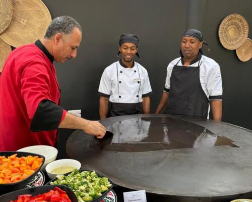 Chef in red preparing a large griddle surrounded by fresh vegetables and sauces, with two cooks watching in a rustic kitchen