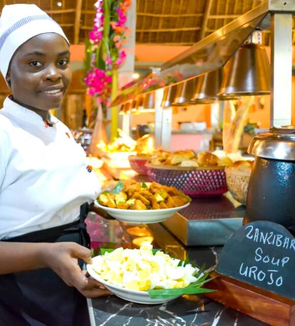 Chef in white uniform serving traditional Zanzibar Urojo soup with egg and fried snacks at a warmly lit buffet in a thatched-roof restaurant