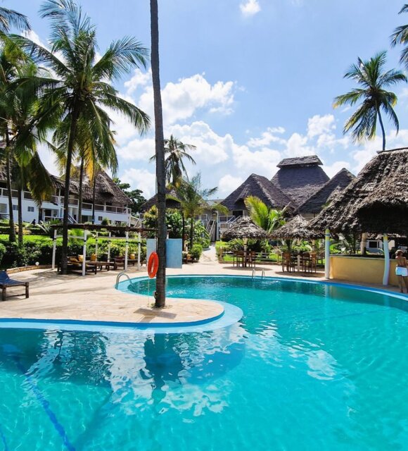 Resort pool surrounded by palm trees, thatched-roof bar, outdoor seating, and bungalow-style accommodations under a sunny blue sky