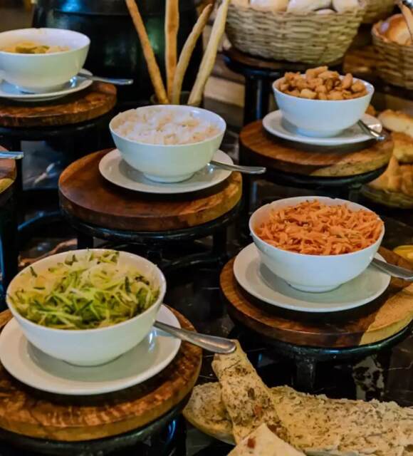 Buffet display with bowls of assorted salads and sides, breadsticks in glass, and woven baskets filled with bread