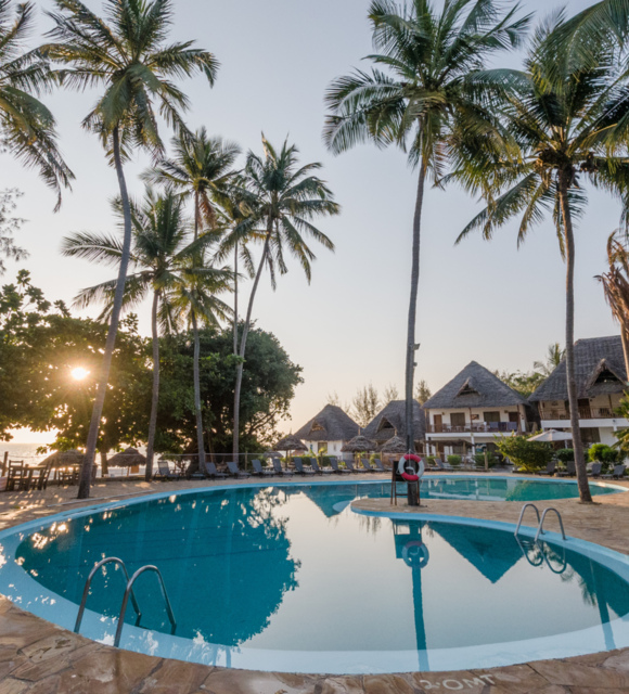 Curved outdoor pool surrounded by palm trees and thatched-roof bungalows at sunset near a beachside resort