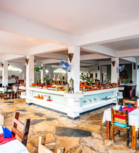 Bright open-air hotel restaurant with rustic wooden chairs, red and blue table settings, and a central buffet with traditional decor