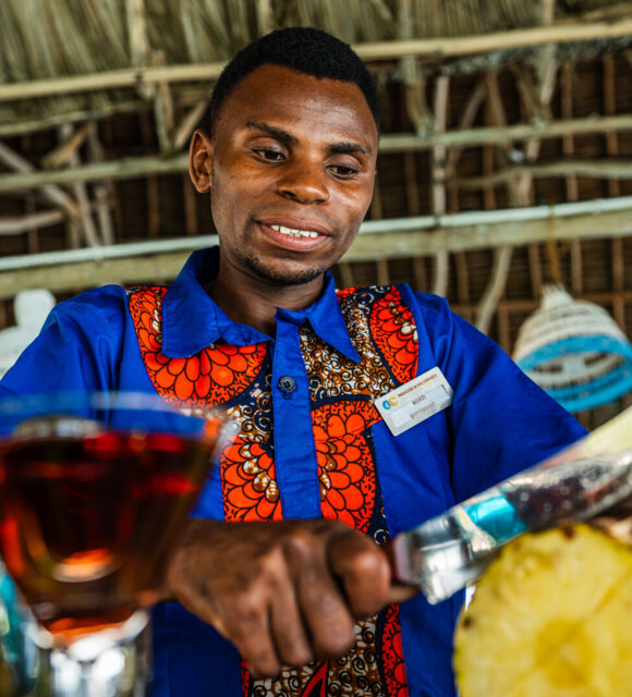 Bartender in a vibrant blue and orange uniform slicing fresh pineapple at a tropical resort bar with thatched roof ceiling