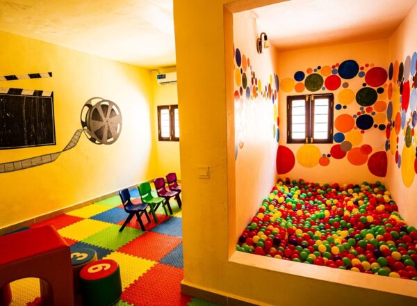Bright children's playroom with colorful foam floor mats, small chairs, a ball pit, and playful wall art featuring film reels and circles
