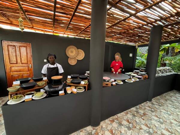 Outdoor hotel buffet area with chefs preparing food under bamboo roof, black counters with dishes, and tropical greenery nearby