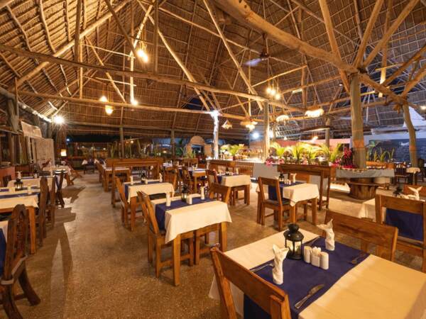 Tropical open-air restaurant with wooden tables, blue and white linens, thatched roof, warm lighting, and tropical plants.