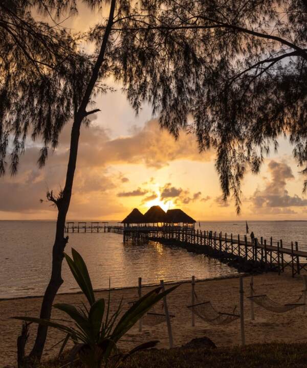 Sunset over a beachfront pier with thatched-roof huts, framed by trees and sandy beach with hammocks for relaxing