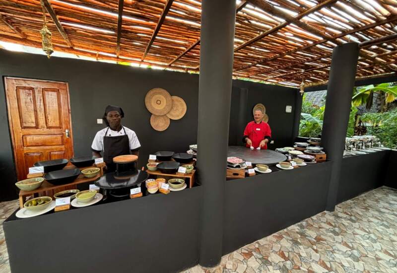 Outdoor hotel buffet area with chefs preparing food under bamboo roof, black counters with dishes, and tropical greenery nearby