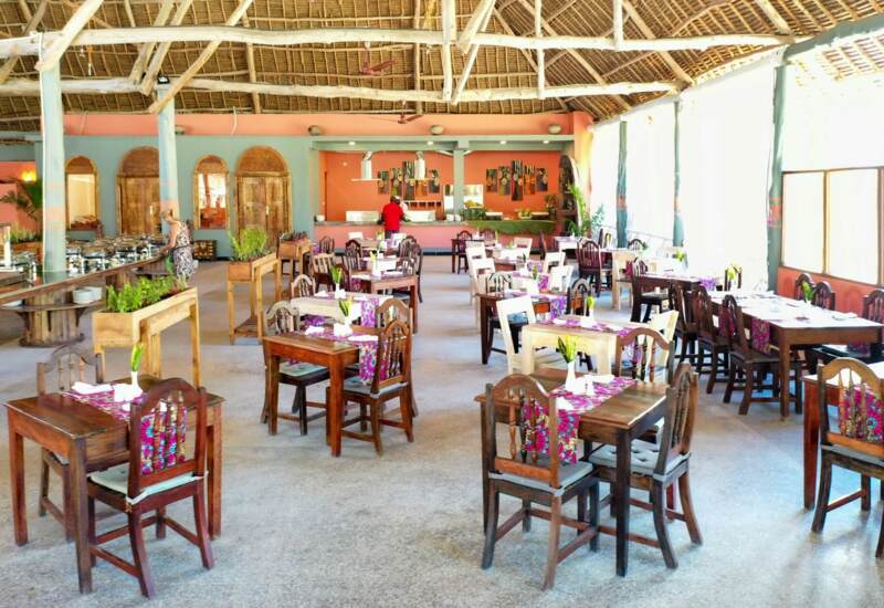 Bright open-air restaurant with wooden furniture, colorful runners, buffet station, and a thatched roof ceiling creating a tropical vibe