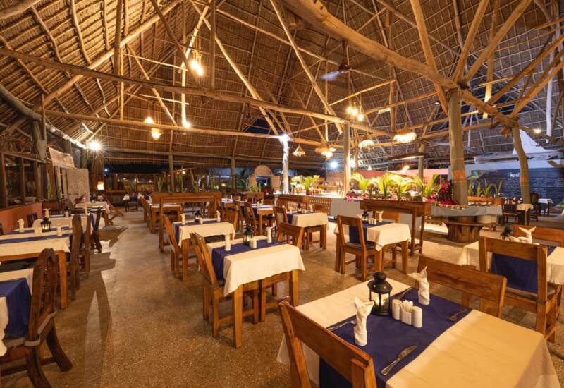 Tropical open-air restaurant with wooden tables, blue and white linens, thatched roof, warm lighting, and tropical plants.