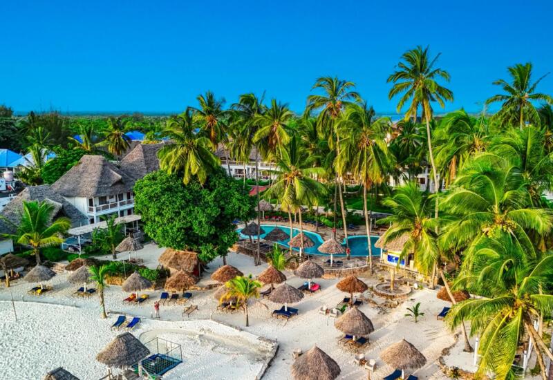 Beachfront resort with thatched roof cottages, palm trees, sandy beach with sun loungers and parasols, and a lagoon pool