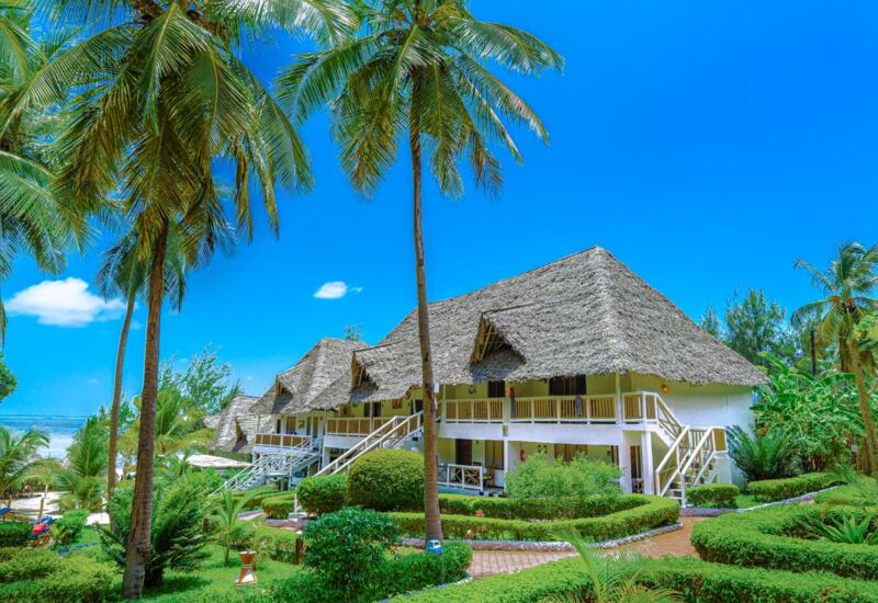 Beachfront resort with thatched-roof cottages surrounded by lush tropical gardens and tall palm trees under a bright blue sky